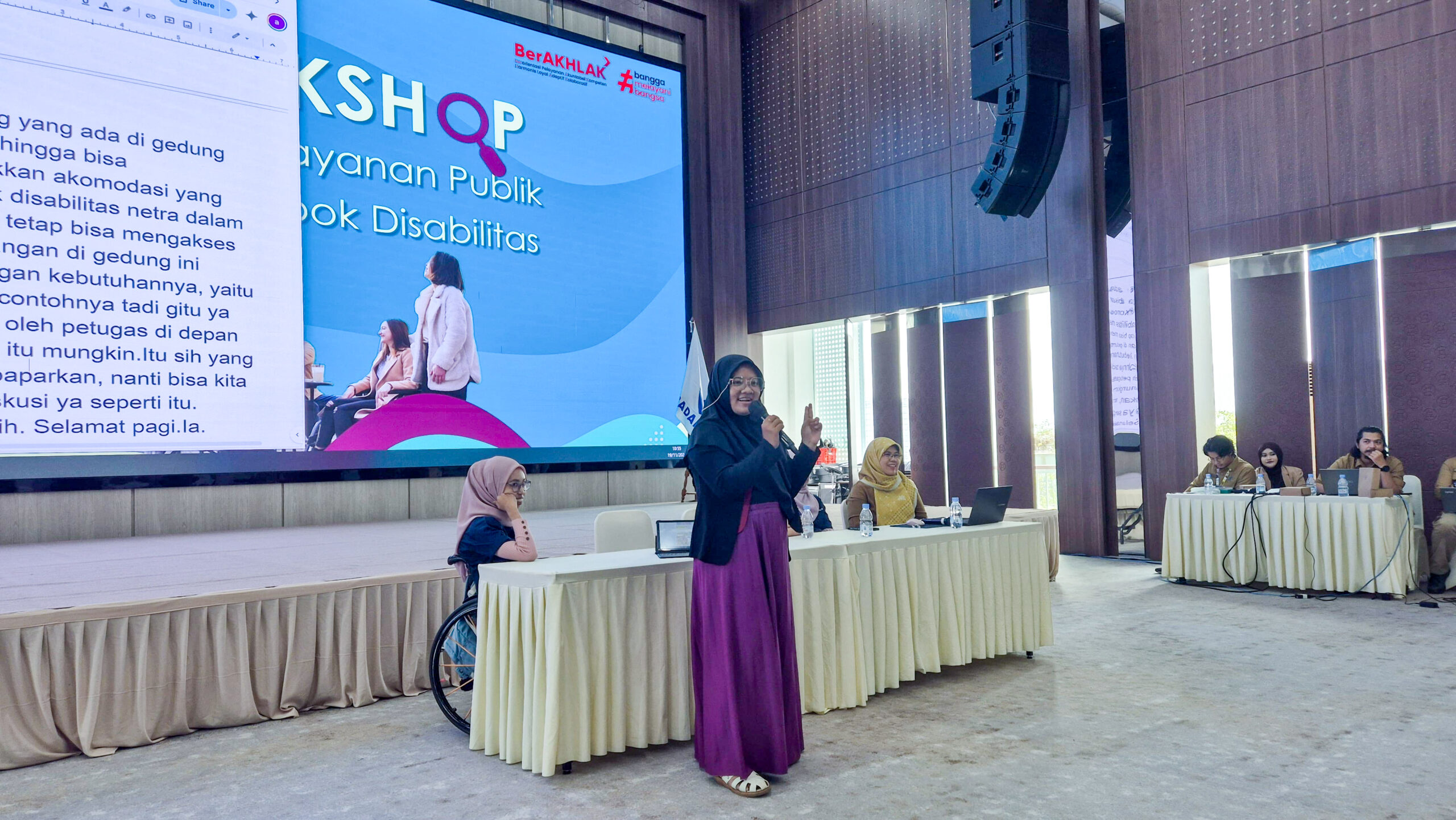 Kinanty Andini stands in front of a long table and speaks into a microphone during a workshop focused on disability inclusion. Behind her, a large screen displays the workshop title in Indonesian. Several panelists, including a woman in a wheelchair, are seated at the front table, while uniformed officials sit to the right.