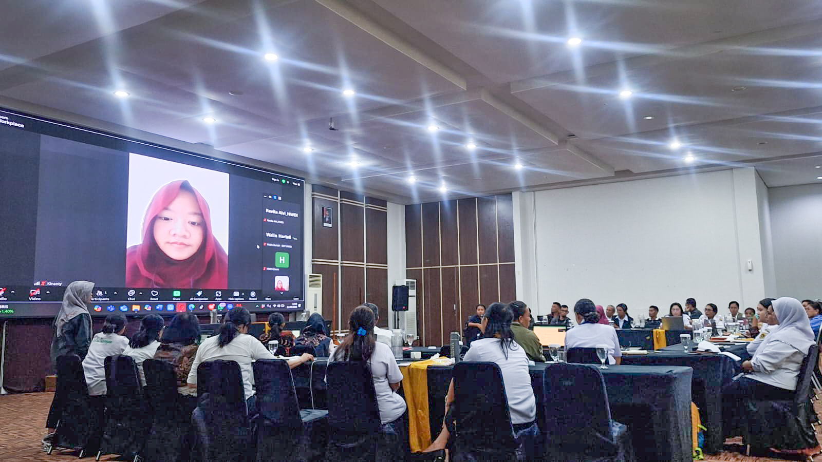 A hybrid training in session with a large group of participants seated in a conference room. On the front wall, a large screen shows Kinanty Andini in a red hijab — addressing the room virtually. Participants are listening attentively, with the room arranged in a U-shape and filled with people of various ages.